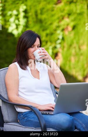 Una donna beve caffè mentre telelavoro con il suo laptop nel giardino della sua casa Foto Stock