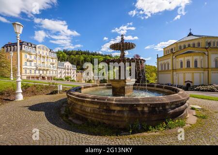 Marianske Lazne, Repubblica Ceca - Maggio 30 2021: Vista della fontana barocca che si erge su piazza Goethe. Edifici, chiesa e alberi in backgroun Foto Stock