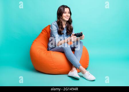 Foto corpo pieno di felice bella giovane donna sguardo vuoto spazio seduta sedia gioco isolato su sfondo colore teal Foto Stock