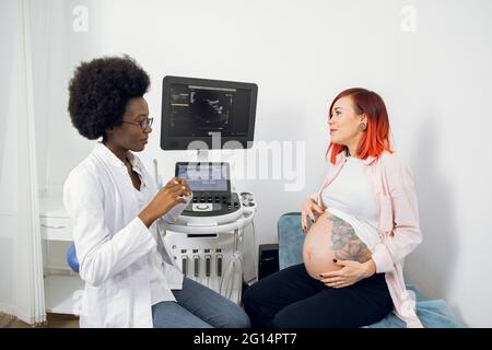 Donna incinta con il bel pancia grande che parla con la sua ostetrica ecografo medico, fiduciosa donna africana in clinica. Terzo controllo del trimestre, gravidanza con gemelli Foto Stock
