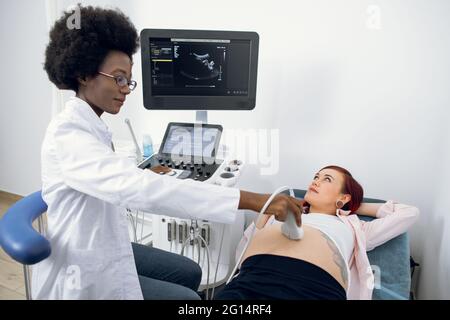 Felice giovane donna caucasica incinta che ha ecografia diagnostica in clinica moderna, guardando la sua donna nera medico ostetrista, mettendo trasduttore a ultrasuoni sul grande cute ventre Foto Stock