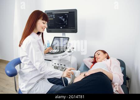 Femmina felice sorridente medico esegue l'esame ecografico della sua paziente femminile per lo screening regolare della gravidanza presso il moderno ufficio medico Foto Stock