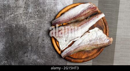 Filetto di pesce di Pollock crudo (nasello) su un tagliere di legno rotondo su sfondo grigio scuro. Vista dall'alto, disposizione piatta Foto Stock