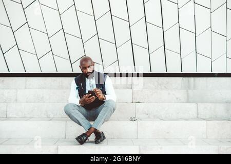 Un imprenditore nero calvo alla moda con una barba e in un abito elegante è seduto su un gradino di marmo sulla strada, di fronte a una wa modellato Foto Stock