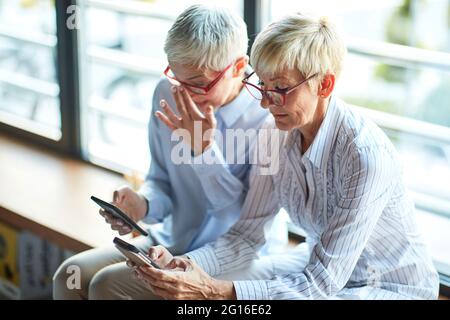 Due donne anziane di aspetto simile che usano smartphone durante la pausa in un'atmosfera rilassata sul posto di lavoro. Affari, ufficio, lavoro Foto Stock