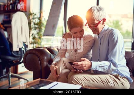 Una donna anziana che mostra un contenuto smartphone alla sua giovane collega che è in pausa dal lavoro in un'atmosfera piacevole in ufficio. Busine Foto Stock