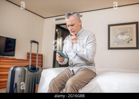 Uomo d'affari di mezza età seduto su un letto in un hotel con uno smartphone nelle mani Foto Stock