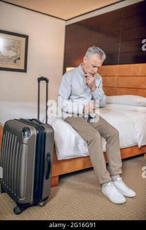 Uomo d'affari di mezza età seduto su un letto in un hotel con uno smartphone nelle mani Foto Stock