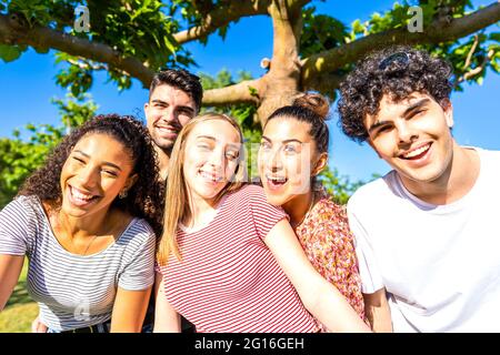 Gruppo di cinque amici molto felici che si divertono al parco in posa guardando la fotocamera per un ritratto. Gen z multirazziale giovani donne e uomini si riunono spensierati Foto Stock