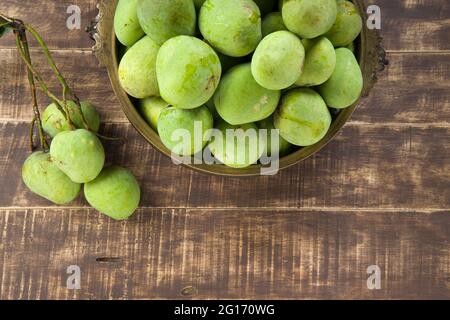 Mango grezzo disposto tradizionalmente in un vaso di ottone e mazzo di mango verde messo vicino su uno sfondo testurizzato di legno. Foto Stock