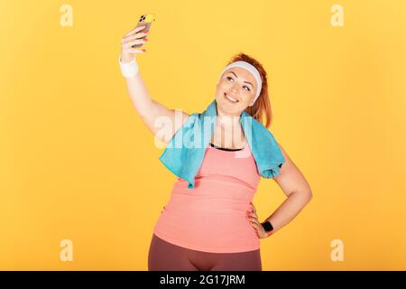 La giovane e gioiosa donna di dimensioni più che indossa abiti sportivi e asciugamano dopo l'allenamento tiene lo smartphone selfie mentre si trova su sfondo giallo. Foto Stock
