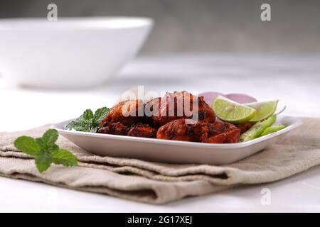 Pollo fritto indiano, sistemato in un piatto bianco guarnito con fette di limone, cipolla, peperoncino verde e foglie di menta che è posto su superficie bianca con gre Foto Stock