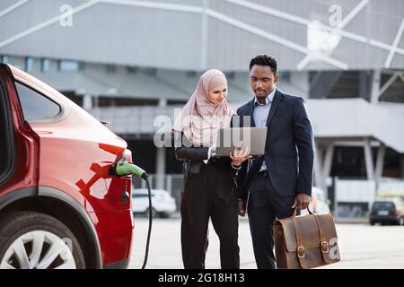 Donna musulmana in hijab che tiene il laptop senza fili e che mostra qualcosa all'uomo africano in costume da lavoro. Uomo e donna in piedi vicino a un'auto elettrica che si sta caricando. Concetto di business auto. Foto Stock