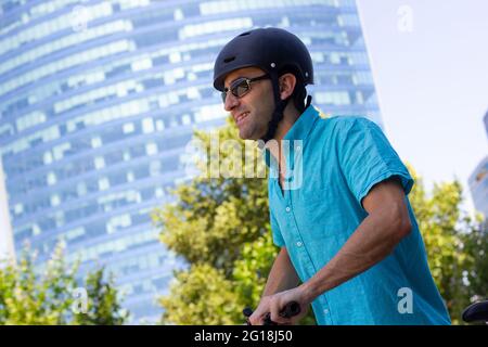 Uomo sorridente con casco in bicicletta in città in giornata di sole. Spostamenti alternativi nell'area di Uban con skyline sullo sfondo Foto Stock