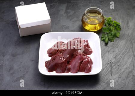 Fegato di pollo crudo disposto splendidamente in una piastra di ceramica bianca con foglia di coriandolo, olio da cucina e scatola di confezionamento su sfondo e posto su t grigio Foto Stock