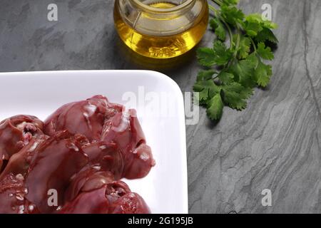 Fegato di pollo crudo disposto splendidamente in una piastra di ceramica bianca con foglie di coriandolo e olio da cucina posto su fondo grigio testurizzato Foto Stock