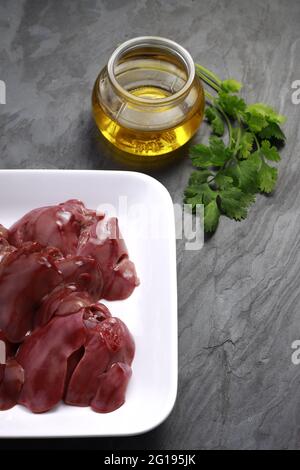 Fegato di pollo crudo disposto splendidamente in una piastra di ceramica bianca con foglie di coriandolo e olio da cucina posto su fondo grigio testurizzato Foto Stock