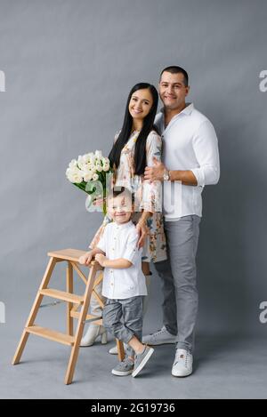 Elegante giovane famiglia: Mamma, papà e figlio di quattro anni si trovano vicino a una scala di legno e sorriso, isolati su uno sfondo bianco. Buona famiglia Foto Stock