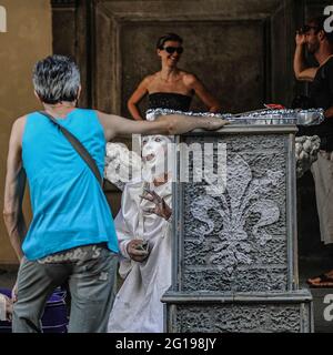 Sbattendo ‘angelo’ con ali diffuse, un volto bianco e guance rosa pallido si stacca la lingua e i gesti mentre si parla con un altro Street performer mentre si fa una pausa dal divertimento dei turisti sulle scale della Galleria degli Uffizi in Piazzale degli Uffizi, Firenze, Toscana, Italia. Numerosi pittori, ritrattisti, caricaturisti, artisti mimi, statue viventi e altri artisti sono autorizzati dalle autorità comunali di Firenze a vendere il loro commercio da piazzole al di fuori delle principali attrazioni culturali della città. Foto Stock