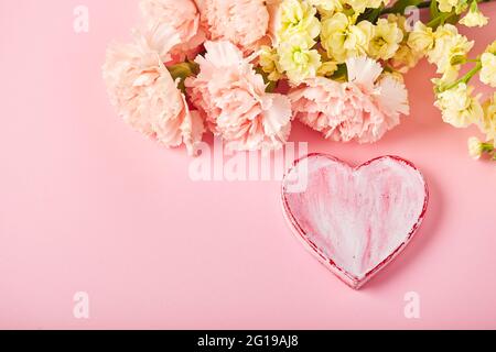 Bouquet di garofani rosa e mattiola gialla e cuore decorativo. Design concetto di saluto per le vacanze con bouquet di garofano su sfondo rosa. Foto Stock