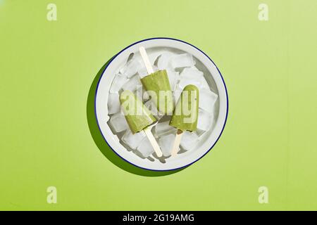 Pop-sicle di pistacchio in casa surgelato in ciotola di ghiaccio su sfondo verde. Papsicle rinfrescante, succo verde congelato sul bastone. Vista dall'alto, spazio di copia. Foto Stock