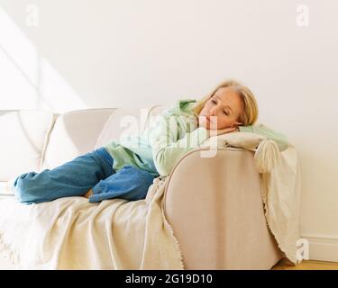 Donna Sad Senior 60 in abiti casual che riposano sul divano in soggiorno a casa, sentendosi depresso durante il pensionamento. Sconvolto donna sola pensò sdraiata sul divano e guardandosi via con tristemente sguardo Foto Stock