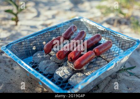 Picnic sulla spiaggia, grigliando le salsicce su una griglia a carbone usa e getta Foto Stock