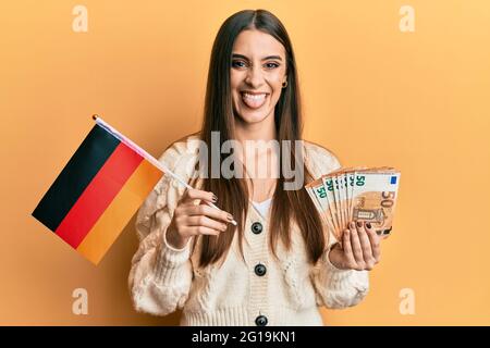 Bella bruna giovane donna che tiene la bandiera della germania e le banconote di euro che attaccano la lingua fuori felice con l'espressione divertente. Foto Stock