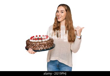 Giovane donna bionda festeggiando il compleanno tenendo grande torta di cioccolato che punta il pollice fino al lato sorridente felice con la bocca aperta Foto Stock