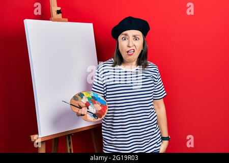 Media età donna ispanica in piedi disegno con tavolozza da pittore cavalletto in faccia d'urto, guardando scettico e sarcastico, sorpreso con mou aperto Foto Stock