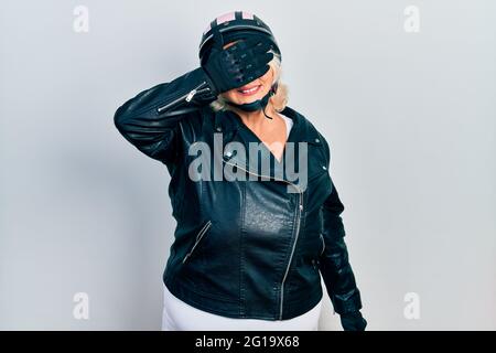 Donna bionda di mezza età che tiene il casco del motociclo sorridente e ridente con la mano sulla faccia che copre gli occhi per sorpresa. Concetto cieco. Foto Stock