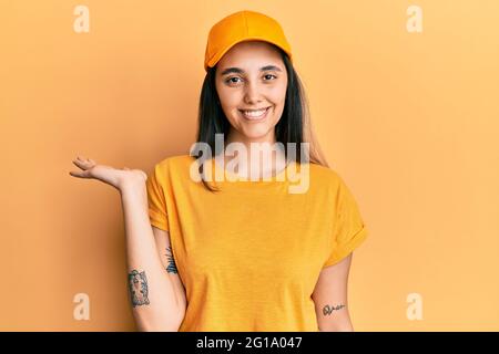 Giovane donna ispanica con uniforme da consegna e cappuccio sorridente allegro presentazione e puntamento con palmo di mano guardando la fotocamera. Foto Stock