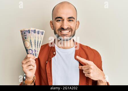 Giovane ispanico che tiene 5000 yen giapponesi banconote sorridendo felice indicando con mano e dito Foto Stock