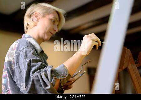 Donna felice dipinge su tela con pennello sottile. La tela si erge sul cavalletto. Artista disegna a easel. Vista laterale della pittura femminile su tela in studio di casa d'arte. Foto di alta qualità. Foto Stock