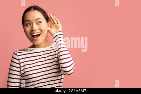 Curiosa donna asiatica allegra che tiene la mano vicino all'orecchio cercando di sentire qualcosa Foto Stock