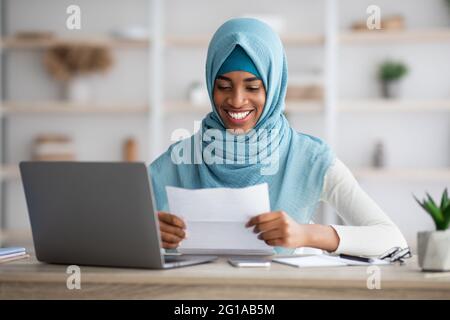 Donna musulmana nera felice in Hijab seduta alla scrivania e lettura della lettera Foto Stock