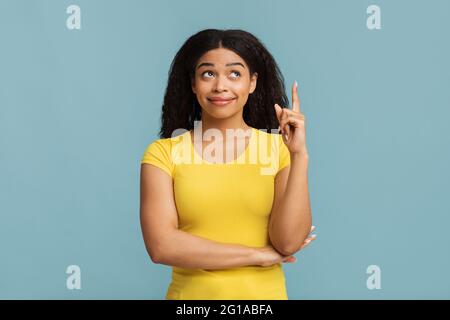 Signora nera che punta il dito verso l'alto e guarda verso l'alto, avendo idea, trovato soluzione, in piedi su sfondo blu Foto Stock