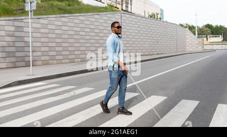 Giovane uomo nero per non vedenti che indossa occhiali scuri, che attraversa la strada della città, utilizzando la canna Foto Stock