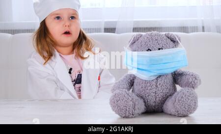 la bambina gioca un medico, fa un'iniezione. Felice bambino medico piccolo esamina un orsacchiotto. Concetto di trattamento del virus corona, iniezione durante Foto Stock