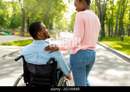 Giovane donna nera e il suo ragazzo disabile in sedia a rotelle trascorrere il tempo insieme all'aperto, vista posteriore Foto Stock