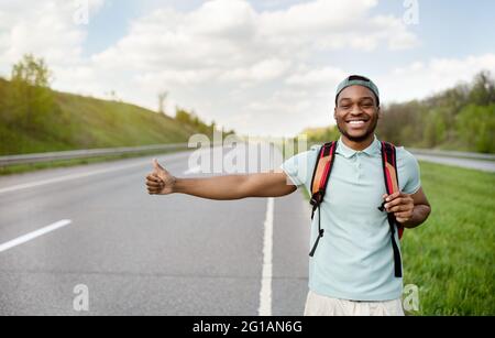 Ragazzo nero sorridente con zaino che si accavallano giù l'automobile, che cattura il giro libero, hitchhiking sull'autostrada, spazio libero Foto Stock