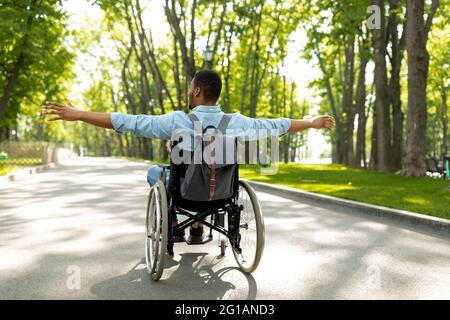Vista posteriore di un giovane uomo nero disabile in sedia a rotelle che si allarga le braccia, sentendosi felice e libero a piedi al parco della città Foto Stock