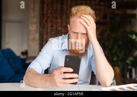 Uomo d'affari redhead confuso in camicia casual intelligente fissando lo schermo dello smartphone, un ragazzo sta leggendo cattive notizie, shock e delusione uomo guarda il telefono con vergogna faccia Foto Stock