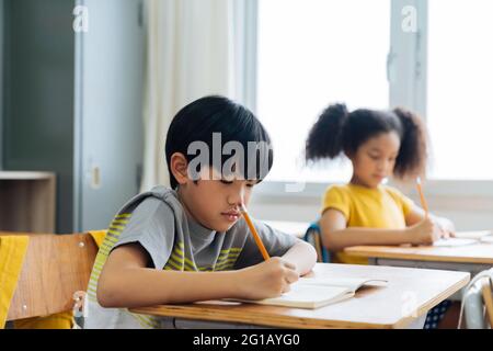 I bambini della scuola seduti alla scrivania in scrittura della scuola in libro di nota con la matita, lo studio, l'istruzione, l'apprendimento. Studenti di sesso maschile e femminile in classe. Ragazzo e ragazza in età elementare Foto Stock