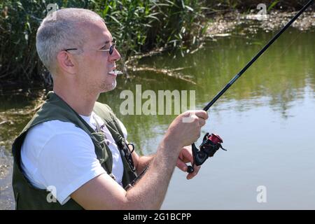 Un pescatore con la sigaretta in bocca e bastone da pesca in mani godendo bella giornata sul lago. Foto Stock
