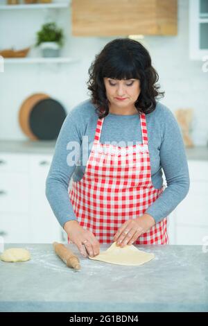 Una donna piuttosto matura stira fuori l'impasto scolpito con un mattarello in cucina sul tavolo. Nonna casalinga. Foto Stock