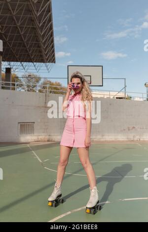 Femmina a piena lunghezza in forma gioiosa con pattina rosa su rulli per scattare foto istantanee su un terreno sportivo soleggiato guardando la fotocamera Foto Stock