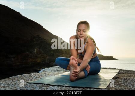 Piano terra di giovani donne che allungano le gambe e la schiena mentre si pratica yoga sulla costa oceanica sotto il sole Foto Stock