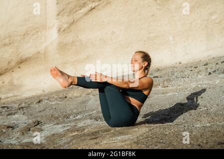 Vista laterale di giovane donna a piedi nudi in abbigliamento sportivo seduto in Ardha Navasana posa mentre pratica yoga alla luce del sole all'aperto Foto Stock