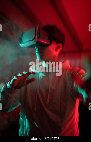 Uomo che indossa una felpa con cappuccio mentre usa occhiali VR e in piedi in studio con luci al neon rosse Foto Stock